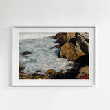 Framed artwork of a coastal scene with rocks and waves on a white wall.